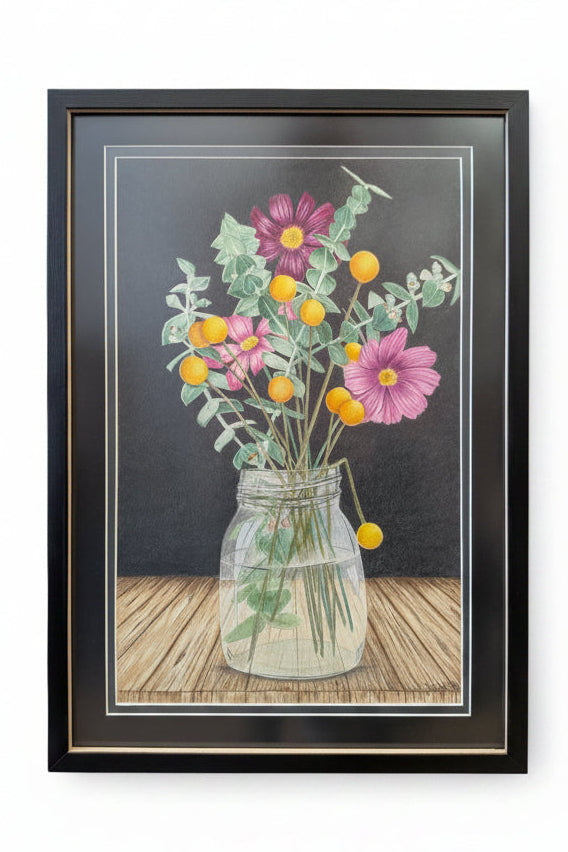 A framed floral artwork featuring pink and purple flowers with yellow-orange blossoms in a clear glass jar, set against a dark background with a wooden tabletop. The artwork is enclosed in a black frame.