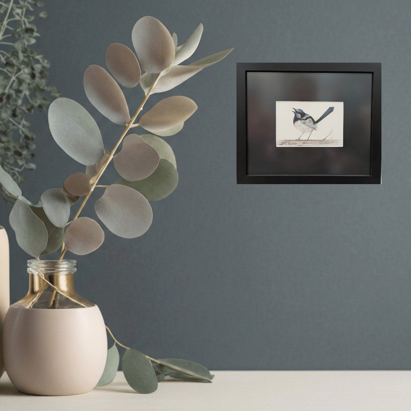 Framed artwork of a blue wren bird on a gray wall with a vase and eucalyptus leaves.